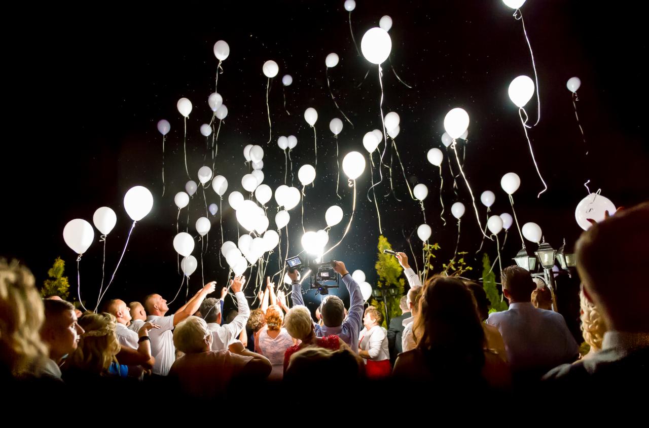 Grupa osób wypuszcza w nocy białe balony, oświetlane przez reflektor kamery, z widocznymi gwiazdami na ciemnym niebie.