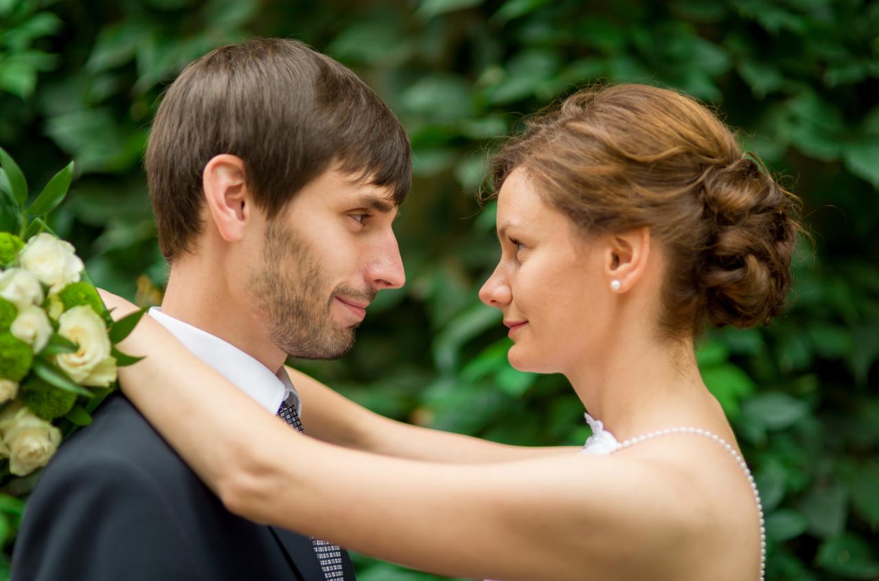 Młoda para, on w ciemnym garniturze z bukietem, ona z upiętymi włosami i perłami, patrzy na siebie na tle zielonych liści.