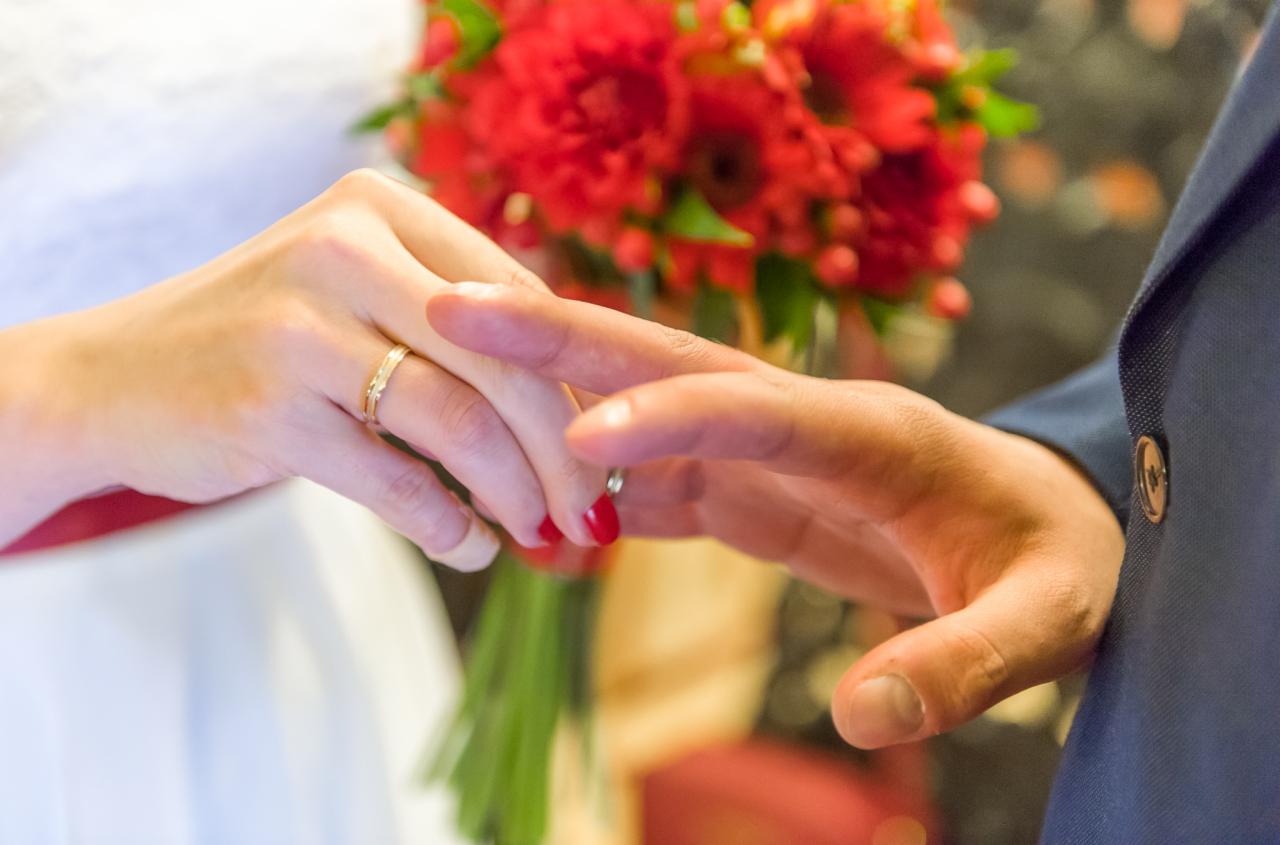 Zbliżenie dłoni podczas ceremonii ślubnej, panna młoda z delikatnym złotym pierścionkiem, pan młody zakłada obrączkę, w tle bukiet czerwonych kwiatów i fragment marynarki.
