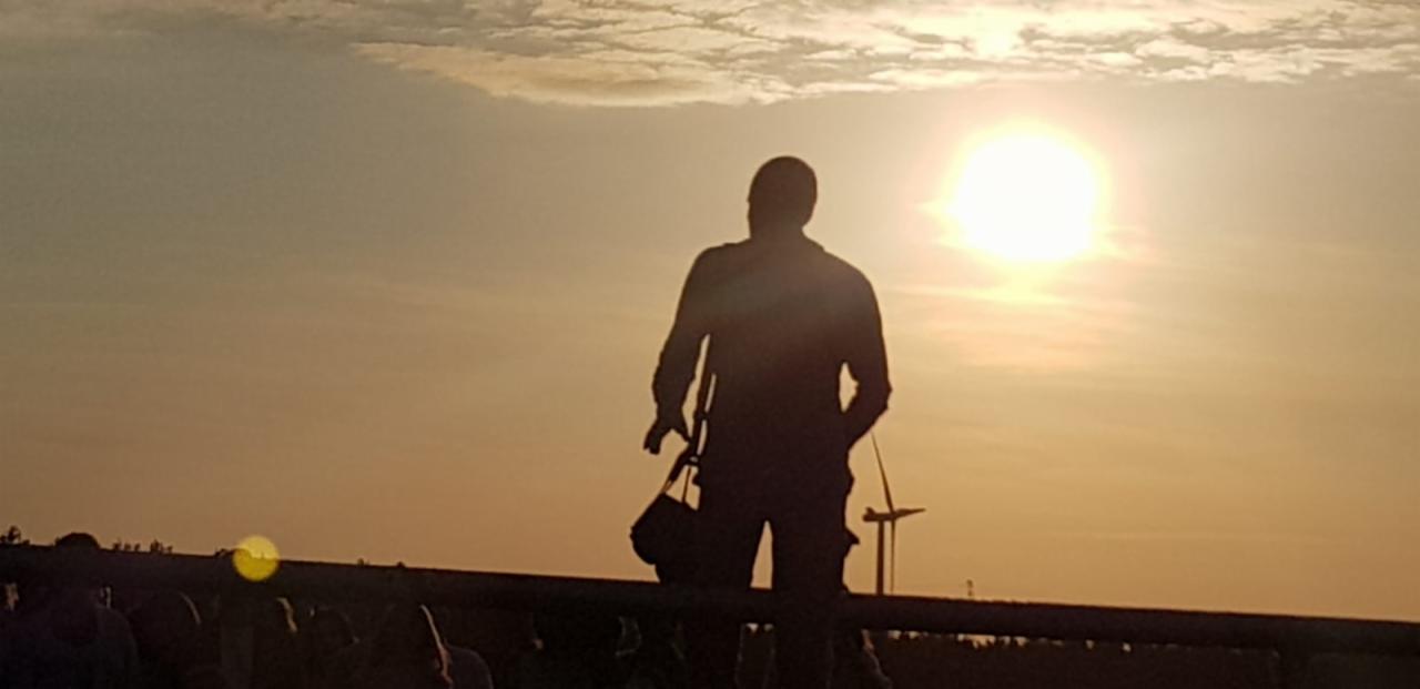 Sylwetka fotografa z torbą na sprzęt na tle zachodzącego słońca i turbin wiatrowych w oddali.