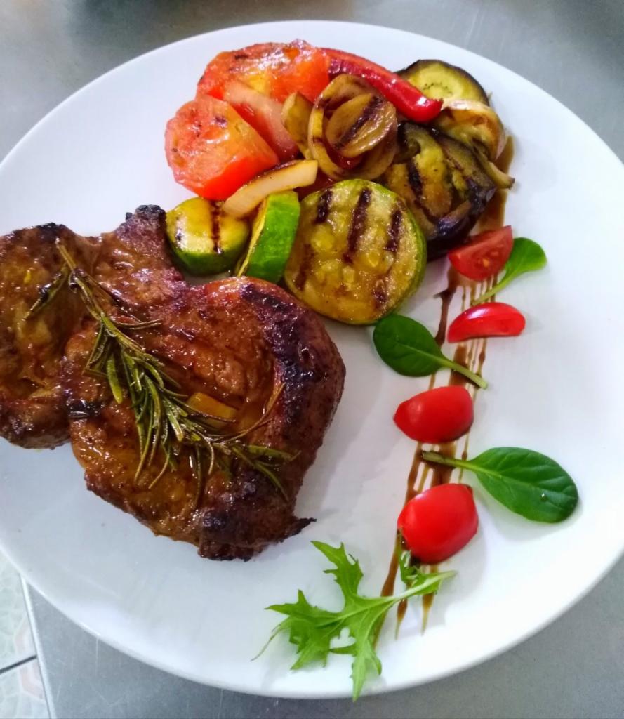 Elegancko podane danie: dwa kawałki mięsa z rozmarynem, grillowane warzywa (cukinia, bakłażan, pomidor, papryka) i dekoracja z pomidorków cherry i listków sałaty, polane sosem balsamicznym...