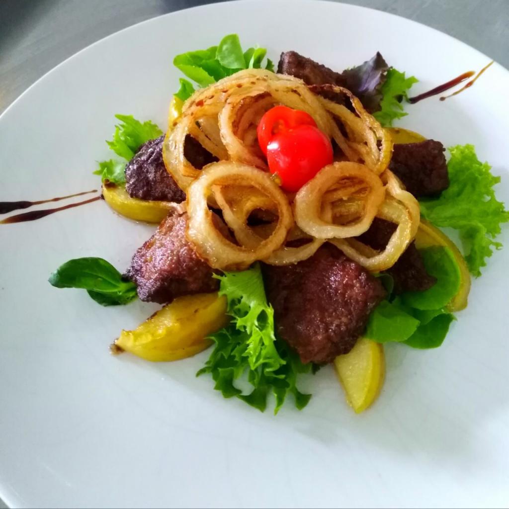 Elegancko podana sałatka z grillowaną wołowiną, krążkami smażonej cebuli, pomidorkiem cherry i sosem balsamicznym na białym talerzu.