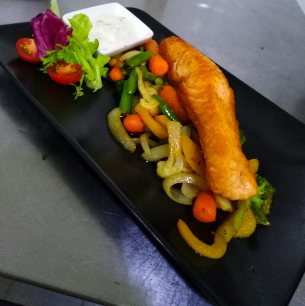 Filet z łososia z grillowanymi warzywami (papryka, cebula, marchewka, brokuły) i sałatką, podany na czarnym, prostokątnym talerzu, z sosem w małej białej miseczce.