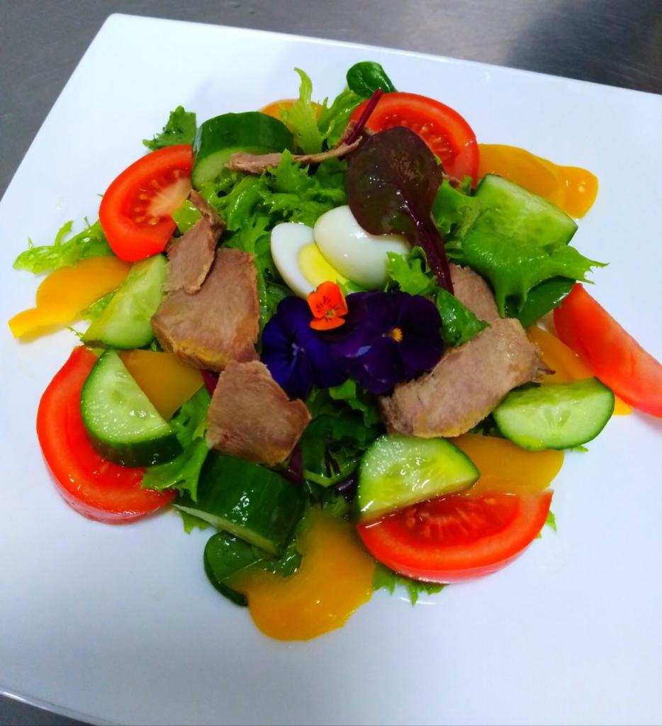 Kolorowa sałatka na białym talerzu z plastrami pomidora, ogórka, żółtej papryki, kawałkami mięsa, jajkiem przepiórczym i jadalnymi kwiatami fiołka.