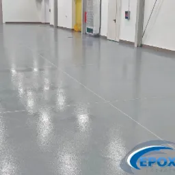 Błyszcząca, szara posadzka w hali przemysłowej z widocznymi liniami podziału i logo firmy w prawym dolnym rogu.