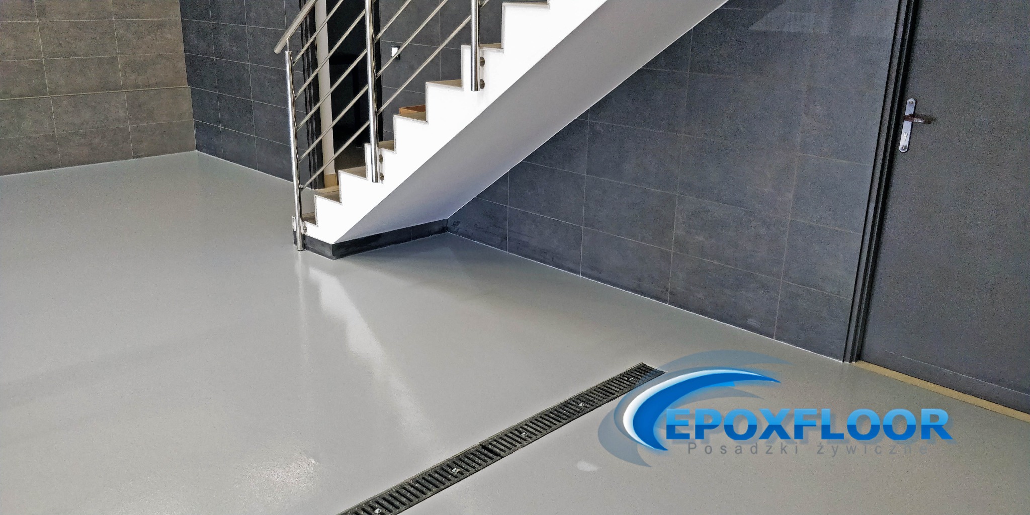 Jasnoszara, gładka posadzka z odpływem liniowym, w tle schody z metalową barierką i ciemnoszare płytki na ścianie, widoczne logo firmy.