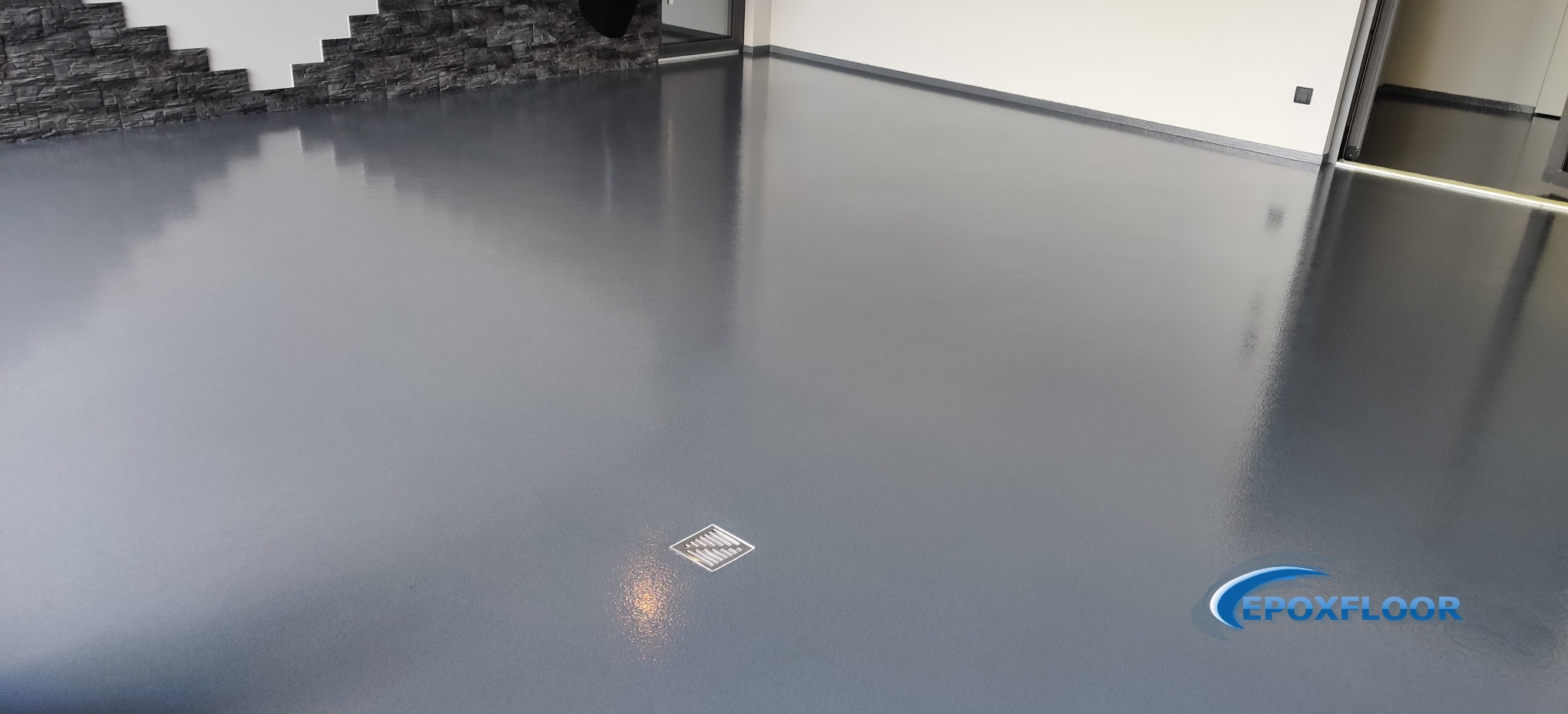 Błyszcząca, szara posadzka żywiczna w pomieszczeniu z fragmentem ściany wykończonej imitacją kamienia, widoczny odpływ liniowy i logo firmy Epoxfloor.