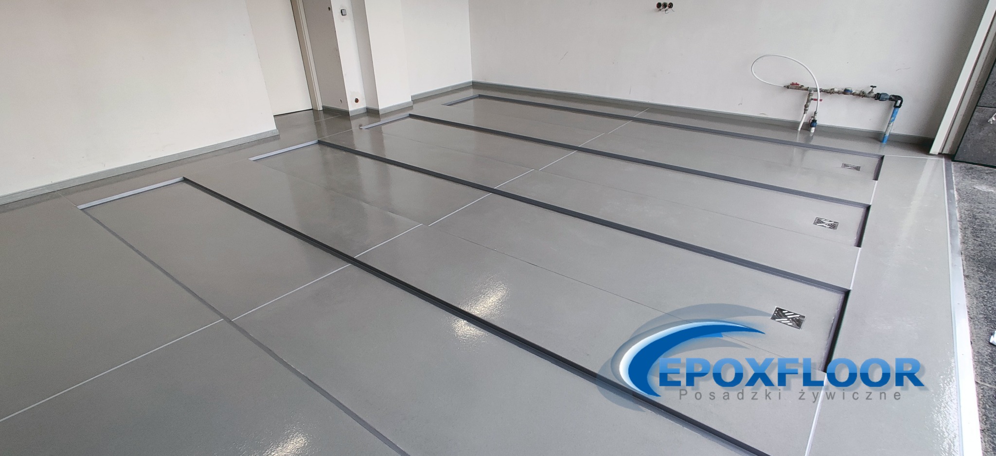 Jasnoszara, błyszcząca podłoga z żywicy w garażu z widocznymi liniowymi odpływami i logo firmy w prawym dolnym rogu.