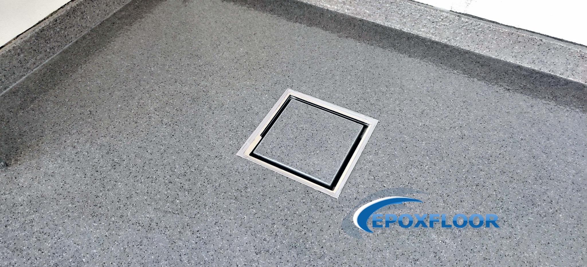 Szara, drobnoziarnista posadzka z odpływem liniowym w kształcie kwadratu, wykończona listwą przyścienną, widoczne logo firmy Epoxfloor.