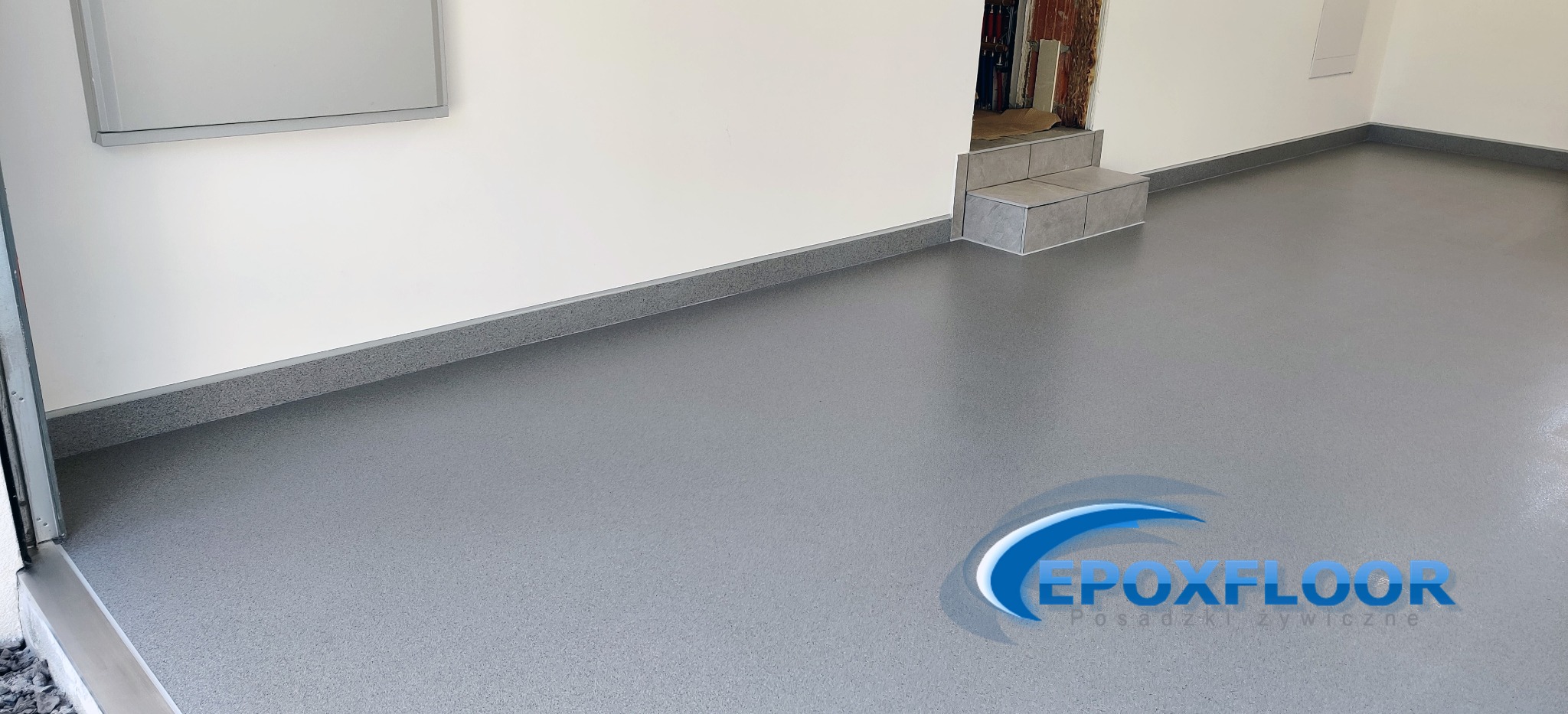 Jasnoszara, jednolita posadzka w garażu lub pomieszczeniu gospodarczym, wykończona listwą przypodłogową w tym samym kolorze, z widocznym logo firmy na podłodze.