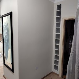 Tomasz Błaszczyk - Ściana w odcieniach szarości przed malowaniem, z widocznym lustrem w czarnej ramie i blokami szklanymi w ścianie przy drzwiach, zabezpieczona folią malarską.