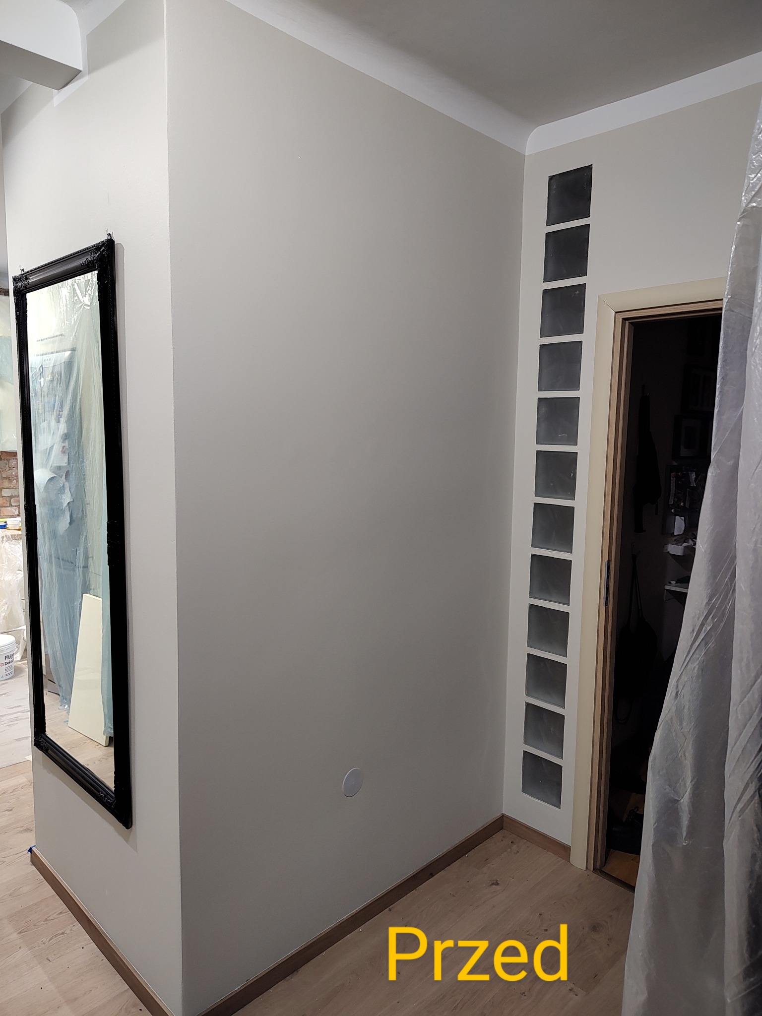 Ściana w odcieniach szarości przed malowaniem, z widocznym lustrem w czarnej ramie i blokami szklanymi w ścianie przy drzwiach, zabezpieczona folią malarską.