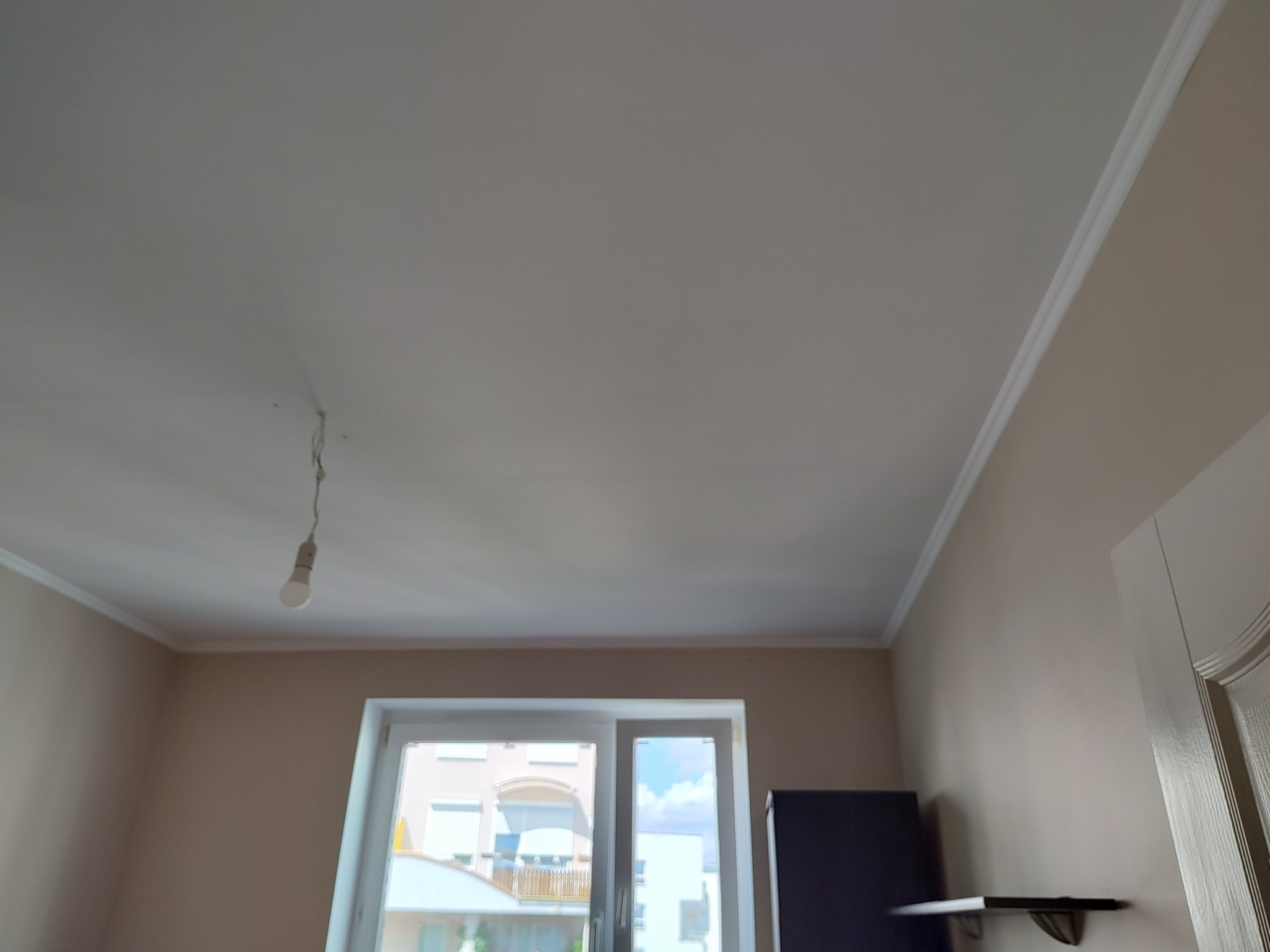 Jasny pokój po malowaniu: gładki, biały sufit z listwami, beżowe ściany, okno z widokiem na budynek. Wisi żarówka bez klosza. Widok z dołu, podkreślający wykonane prace malarskie.