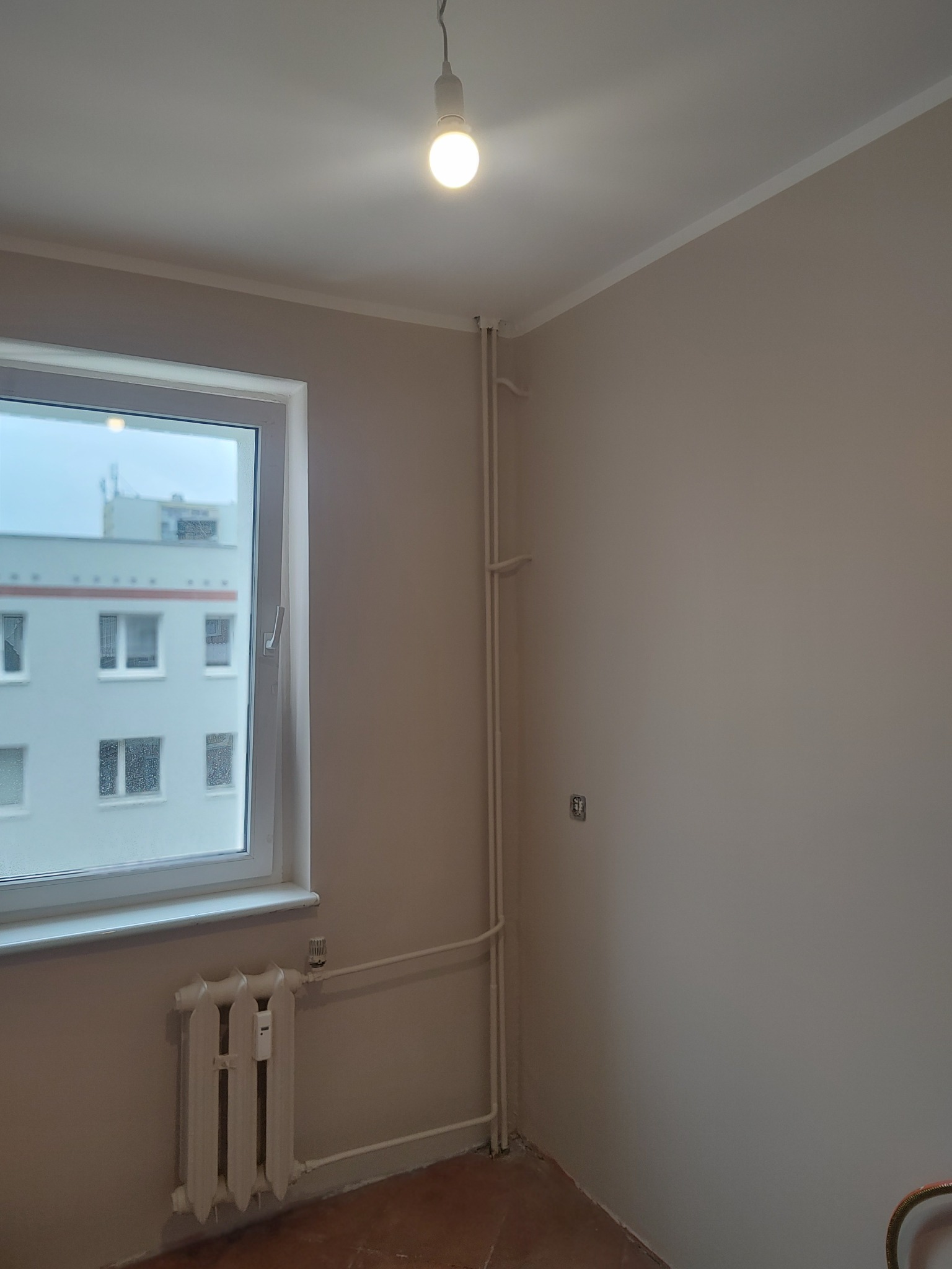 Pomieszczenie po malowaniu ścian na beżowo, widoczne okno, grzejnik i rury. Świecąca żarówka oświetla wnętrze. Wykończone ściany, gotowe do dalszych prac.