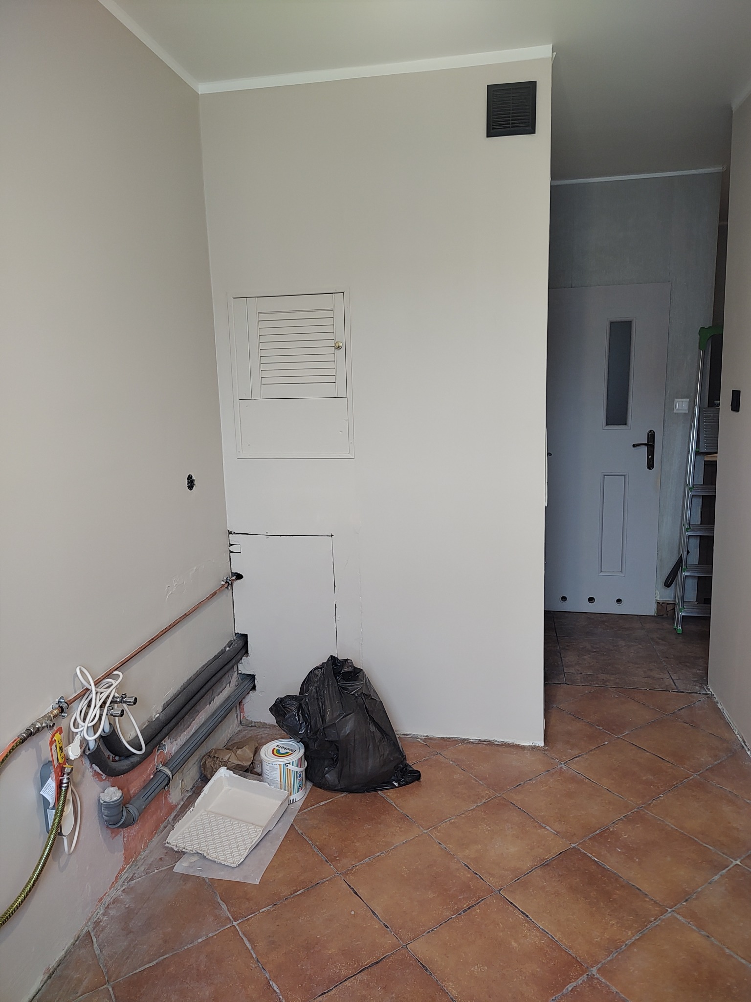 Ściana po malowaniu na jasny kolor, widoczne rury, grzejniki, narzędzia malarskie i fragment podłogi z terakoty. W tle drzwi i drabina.