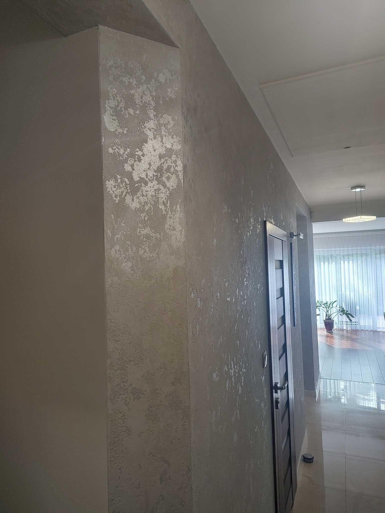 Ściana z dekoracyjną, srebrzystą strukturą w przedpokoju. Widoczna drewniana ościeżnica i fragment wnętrza z rośliną doniczkową.