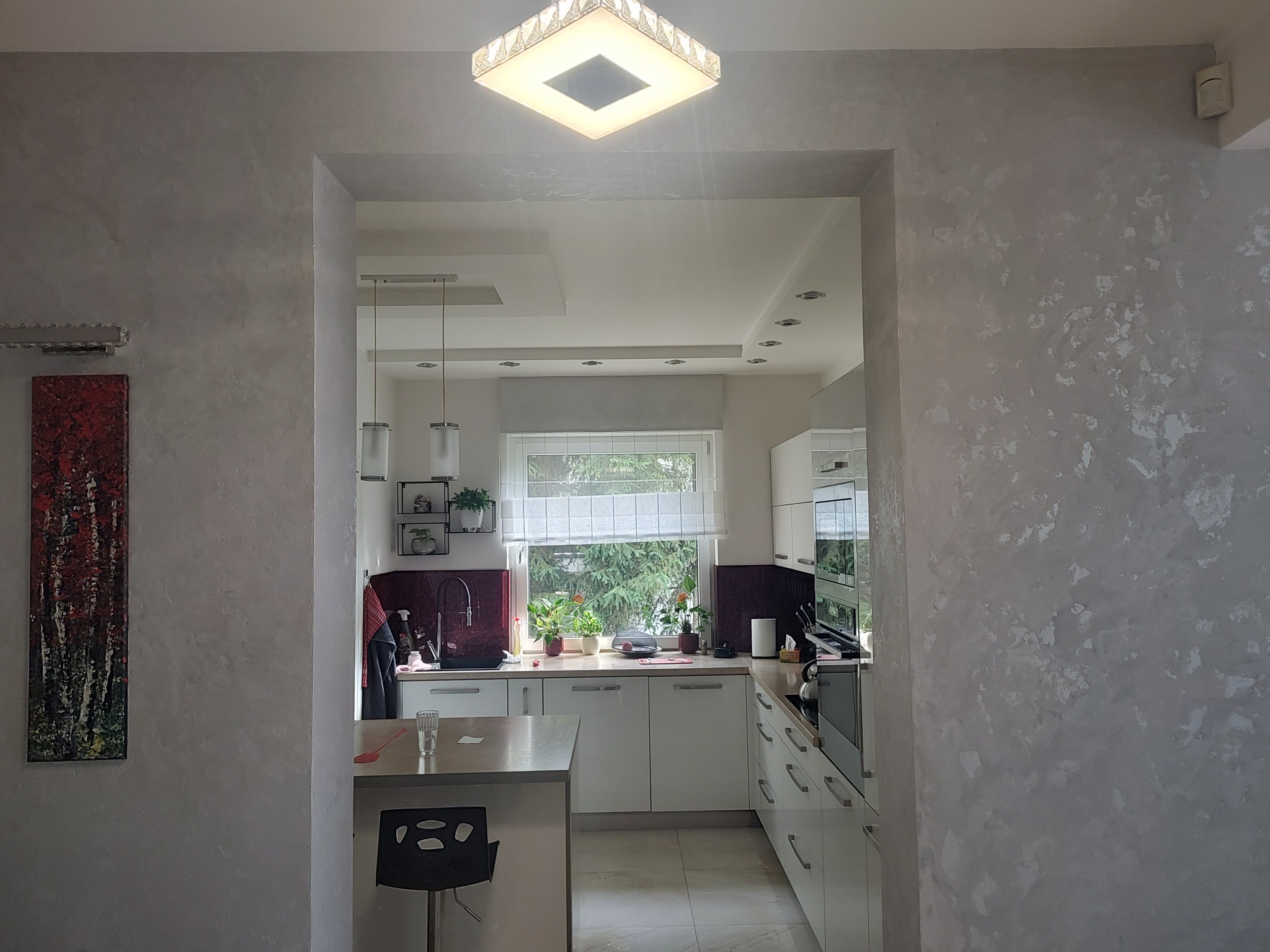 Elegancka kuchnia z białymi meblami widziana przez wykończony dekoracyjną szarą farbą otwór w ścianie. Na ścianie obraz w odcieniach czerwieni i brązu.