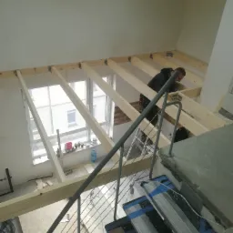 Widok z góry na drewnianą konstrukcję sufitu podwieszanego w trakcie montażu, z pracownikiem w czarnej bluzie w tle, przez metalową barierkę schodów. W tle okno i ściana z częściowo położoną cegłą.