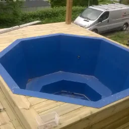 Montaż drewnianego tarasu wokół niebieskiej wanny jacuzzi, w tle srebrny samochód dostawczy z logo firmy budowlanej.