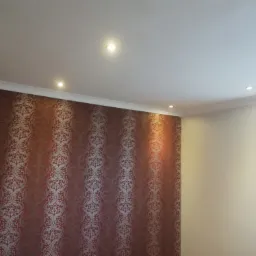 Wnętrze pokoju z dekoracyjną tapetą w odcieniach czerwieni i złota na jednej ścianie, gładką, kremową ścianą z oknem po prawej stronie oraz białym sufitem z wbudowanymi punktowymi lampami LED.