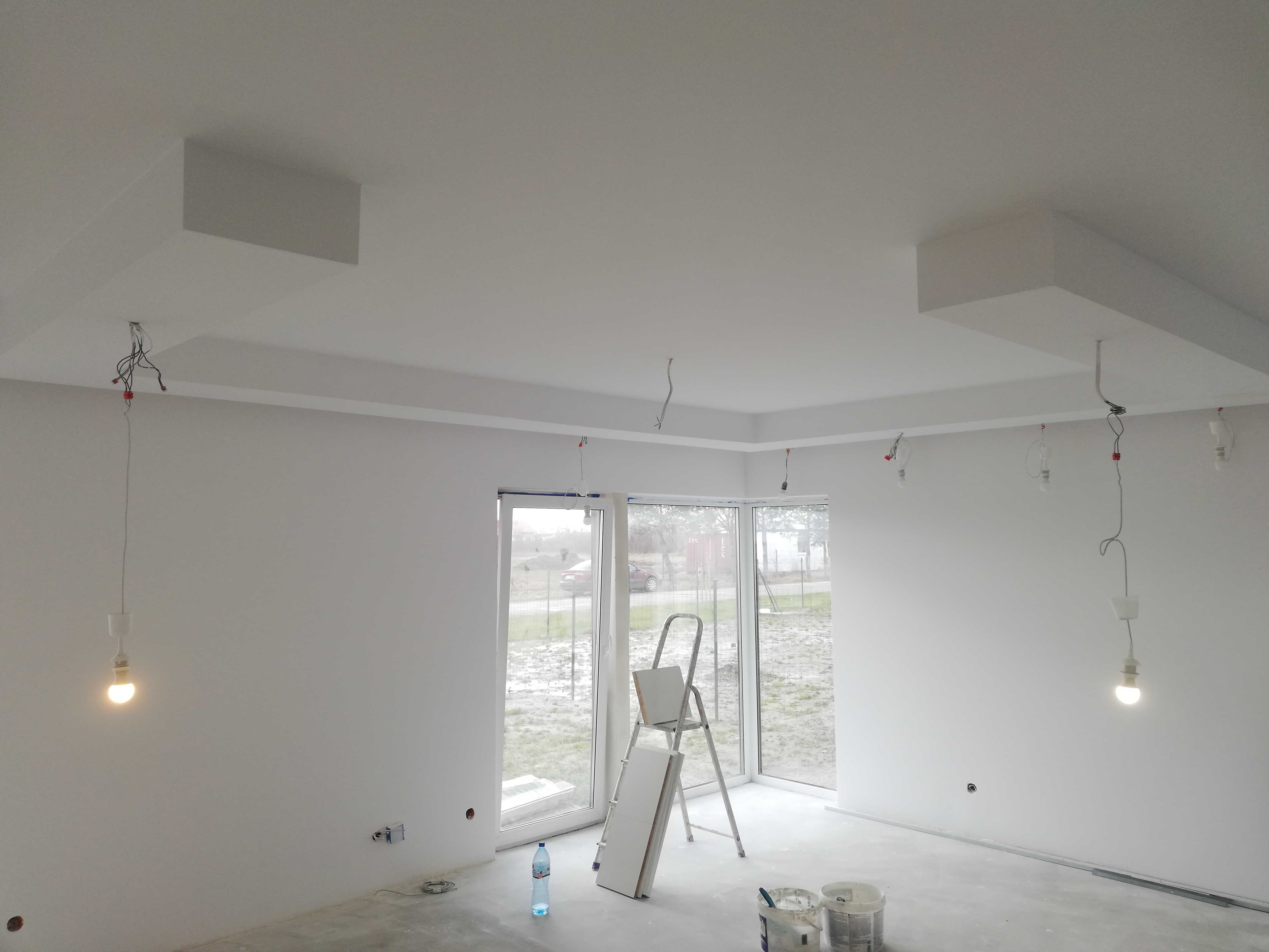 Wnętrze w stanie deweloperskim z przygotowanymi punktami oświetleniowymi, świeżo pomalowanymi ścianami i sufitami, drabina oparta o okno balkonowe, widoczne puszki z farbą i butelka wody na podłodze.