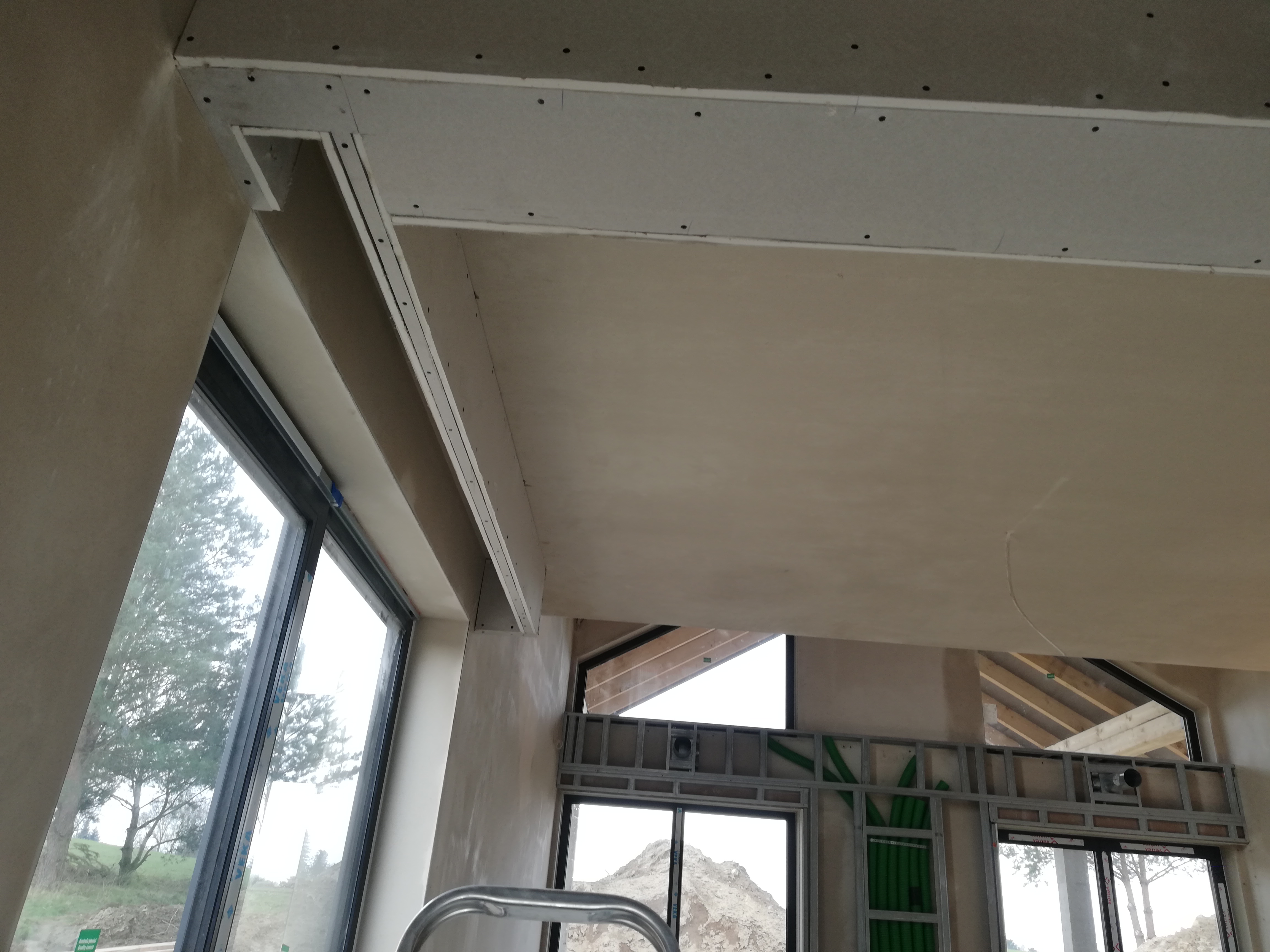 Wnętrze w trakcie prac wykończeniowych: widoczny sufit z zamontowaną płytą kartonowo-gipsową, ściany po nałożeniu gładzi, metalowa konstrukcja pod zabudowę okienną z instalacją elektryczną...