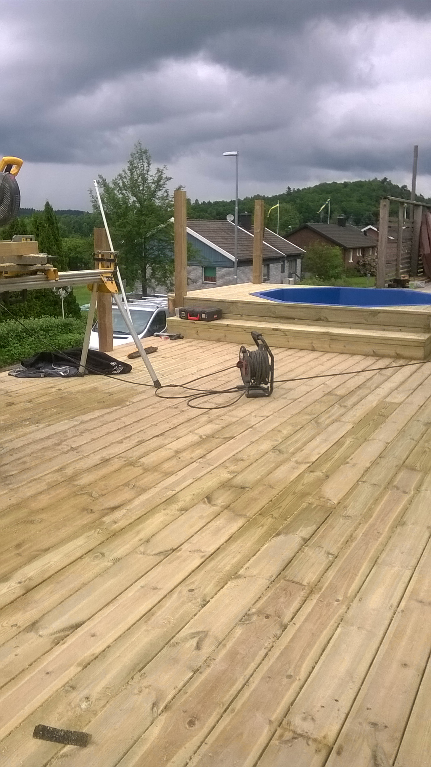 Budowa drewnianego tarasu z wbudowanym basenem, widoczny sprzęt budowlany i narzędzia na tle pochmurnego nieba i zabudowań.