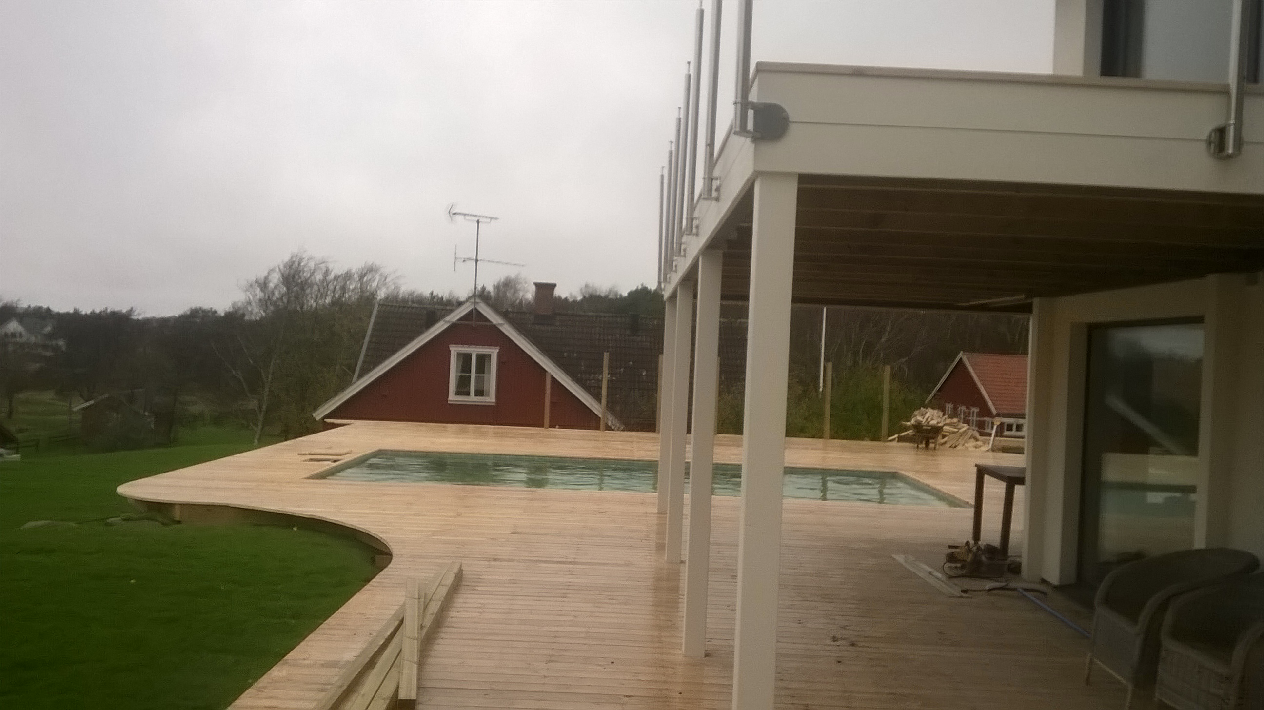 Nowoczesny taras z drewna z dwoma małymi basenami, widok na czerwoną chatę z anteną na dachu w tle, pochmurna pogoda.