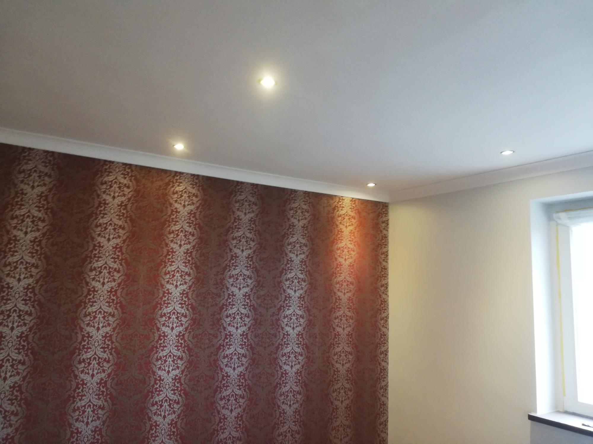 Wnętrze pokoju z dekoracyjną tapetą w odcieniach czerwieni i złota na jednej ścianie, gładką, kremową ścianą z oknem po prawej stronie oraz białym sufitem z wbudowanymi punktowymi lampami LED.
