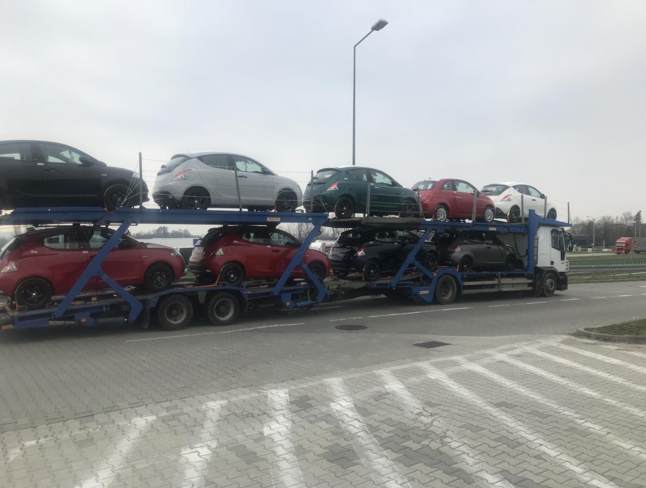 Ciężarówka z lawetą przewożąca osiem samochodów Lancia Ypsilon w różnych kolorach na dwupoziomowej platformie. Widoczne samochody w kolorach: czarnym, szarym, zielonym, czerwonym i białym. Laweta...