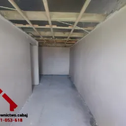 Wnętrze pomieszczenia po tynkowaniu maszynowym, widoczne świeżo nałożone, gładkie tynki na ścianach i surowy strop z drewnianych belek, logo firmy w lewym dolnym rogu.