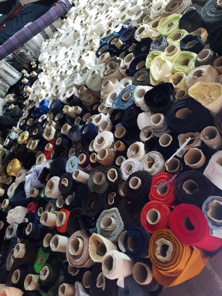 Różnokolorowe rolki tkanin i dzianin ułożone ciasno na półce w magazynie, widoczne różne faktury i odcienie, od jasnych bieli po głębokie granaty i czerwienie.