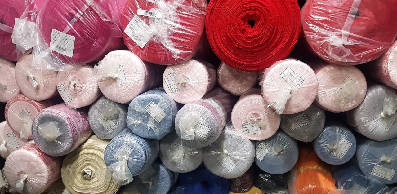 Magazyn z rolowanymi belami tkanin w różnych kolorach, zapakowanymi w przezroczystą folię, z widocznymi etykietami informacyjnymi.