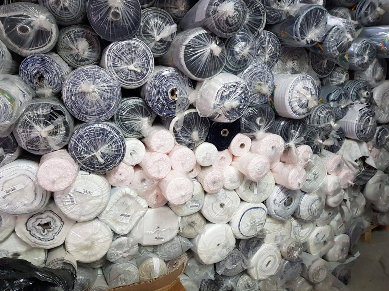 Stos rolek różnokolorowych tkanin i dzianin, zapakowanych w przezroczystą folię, ułożonych warstwami w magazynie hurtowni.