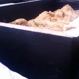 Świeżo zaizolowane fundamenty z czarną powłoką hydroizolacyjną, widoczny wykop z piaskiem i fragment białej folii ochronnej na krawędzi.