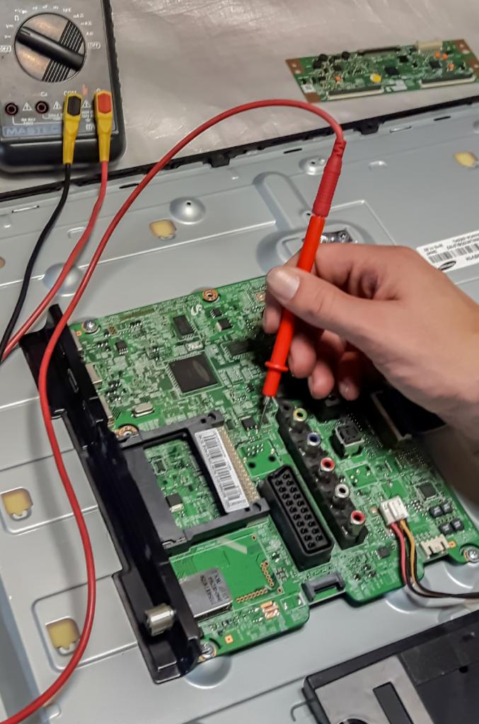 Pomiar napięcia na płycie głównej telewizora za pomocą multimetru, widoczne przewody pomiarowe i fragment obudowy telewizora.