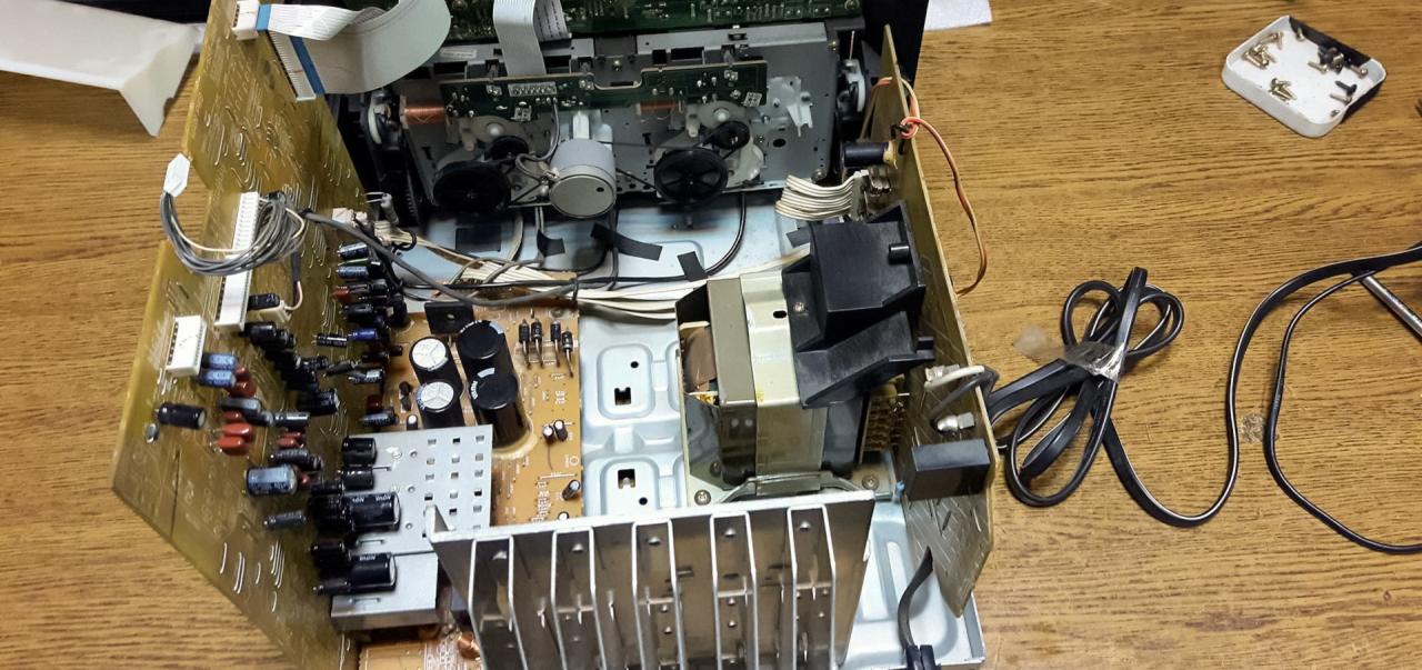 Wnętrze rozmontowanego sprzętu RTV, widoczne płyty główne, transformator, kondensatory i kable na drewnianym blacie.