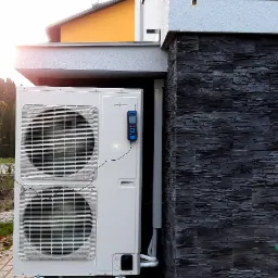 Biała zewnętrzna jednostka pompy ciepła marki Viessmann zamontowana przy ścianie budynku wykończonej czarnym kamieniem dekoracyjnym, z podłączonym urządzeniem diagnostycznym.