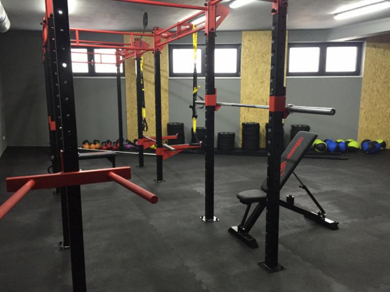 Sala ćwiczeń z czarno-czerwoną konstrukcją do podnoszenia ciężarów, ławeczką z napisem 'CHAMPION' i zestawem kettlebells w tle