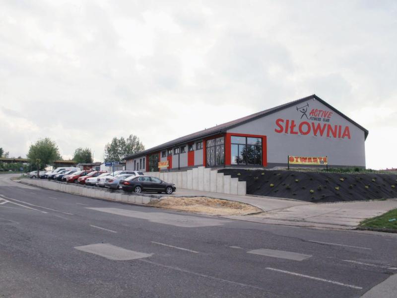 Zewnętrzny widok budynku siłowni Active Fitness Club z dużym napisem 'Siłownia' na elewacji, parkingiem i zagospodarowanym terenem przed wejściem.