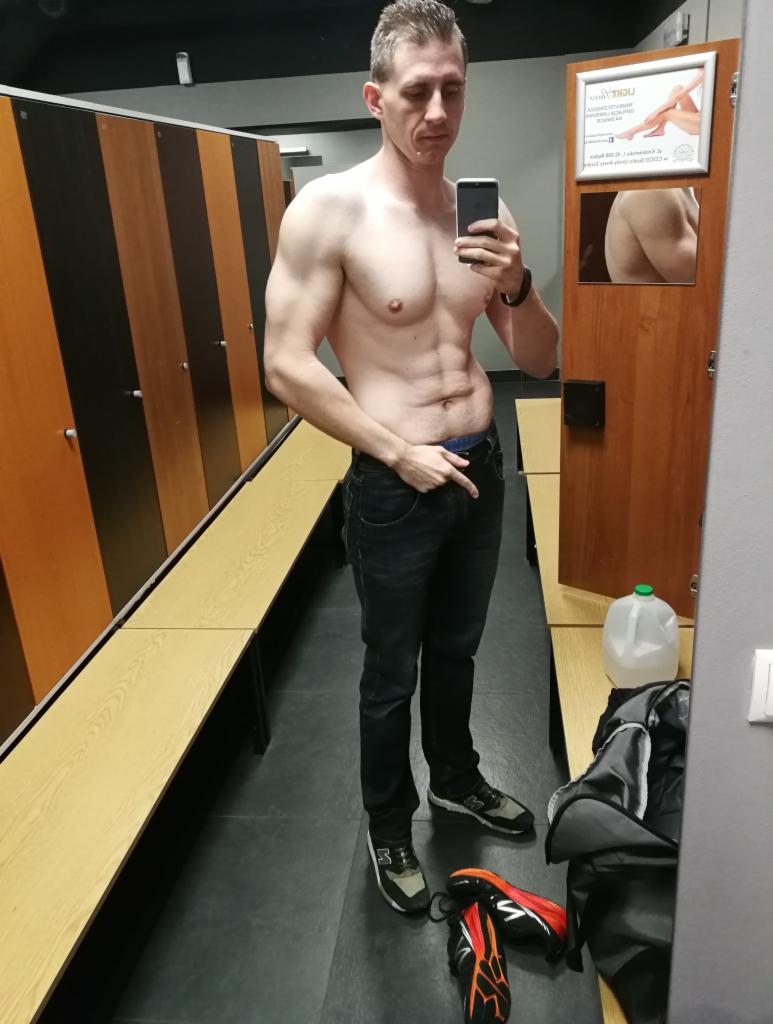 Mężczyzna robi selfie w szatni siłowni, eksponując umięśnioną sylwetkę; widoczny plakat reklamowy na drzwiach w tle.