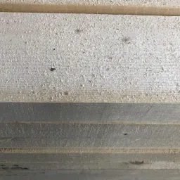 Stos desek z jasnego drewna, pokrytych drobnymi wiórami, widoczne słoje.