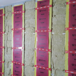 Centrum Dystrybucji ogrzewania i Fotowoltaiki Red Snake - Ściana w trakcie izolacji wełną mineralną, zabezpieczona folią z nadrukiem 'Red Snake' i taśmą mocującą, widoczna metalowa konstrukcja nośna.