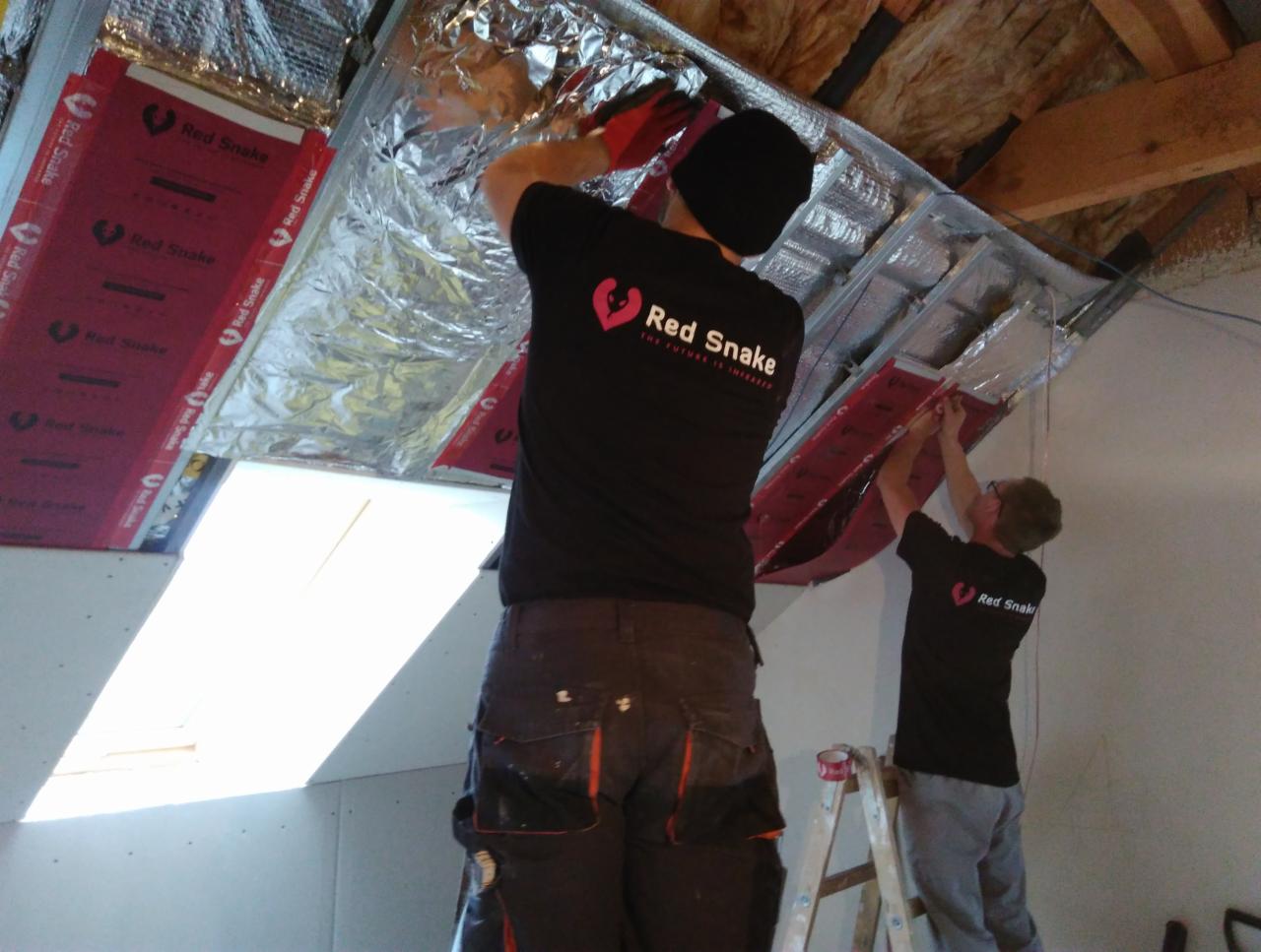 Dwóch pracowników w czarnych koszulkach z logo 'Red Snake' instaluje izolację termiczną na poddaszu, widoczne panele izolacyjne z czerwoną folią i srebrną warstwą odbijającą, jeden stoi na drabinie...