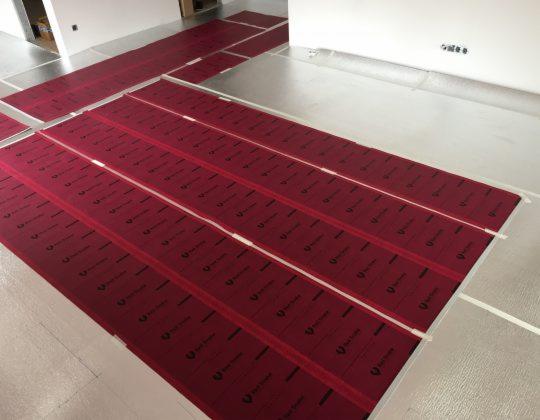 Montaż mat grzewczych: czerwone maty z czarnym nadrukiem układane na srebrnej folii izolacyjnej, zabezpieczone taśmą, widok z góry.