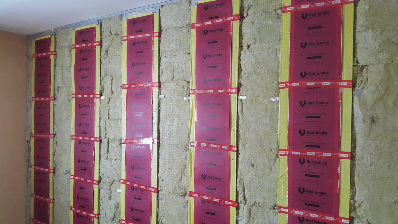Ściana w trakcie izolacji wełną mineralną, zabezpieczona folią z nadrukiem 'Red Snake' i taśmą mocującą, widoczna metalowa konstrukcja nośna.