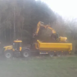 Żółty ciągnik z naczepą i koparka podczas załadunku ziemi, widoczne drzewa w tle, pochmurna pogoda.