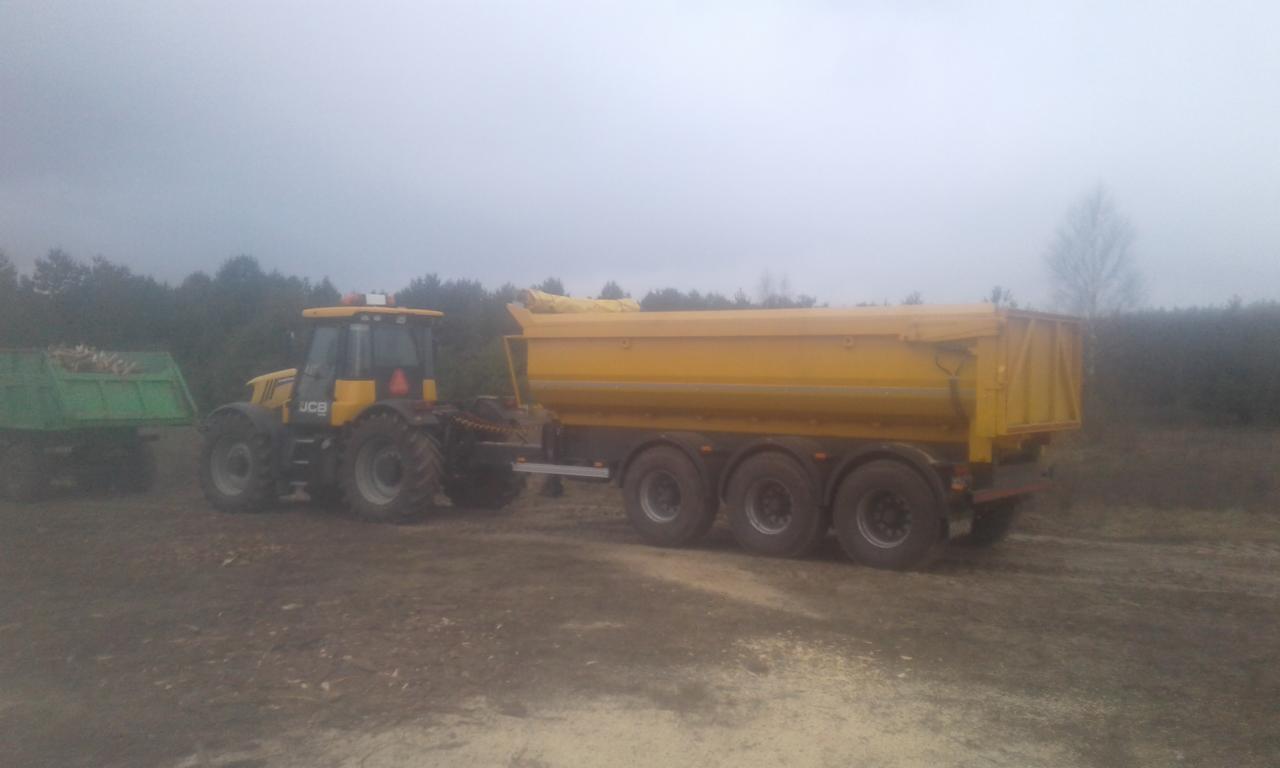Żółty ciągnik JCB z przyczepą wywrotką na grząskim terenie, w tle zalesiony obszar i mglista pogoda. Dodatkowo widoczna mniejsza, zielona przyczepa.