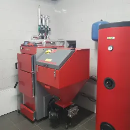 Czerwono-szary piec na paliwo stałe z zasobnikiem i zbiornikiem buforowym w kotłowni wyłożonej białymi płytkami.