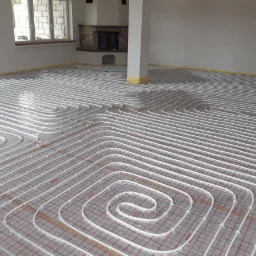 Białe rurki ogrzewania podłogowego ułożone spiralnie na czerwonej siatce izolacyjnej w jasnym, przestronnym pomieszczeniu z kominkiem w tle.